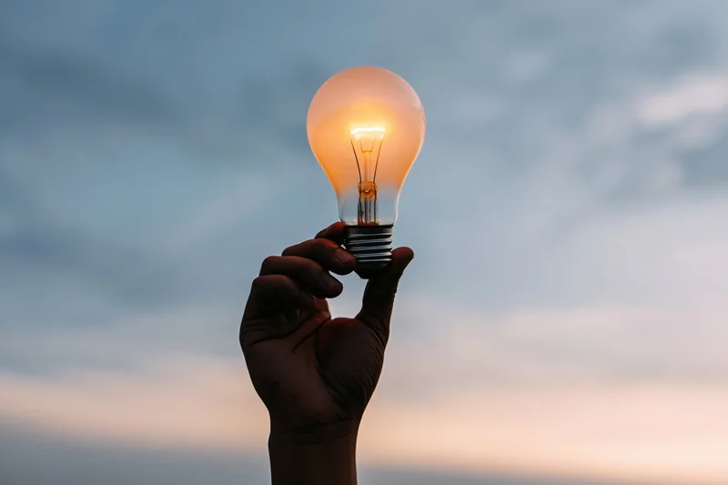 Hand holding glowing incandescent lightbulb against twilight sky