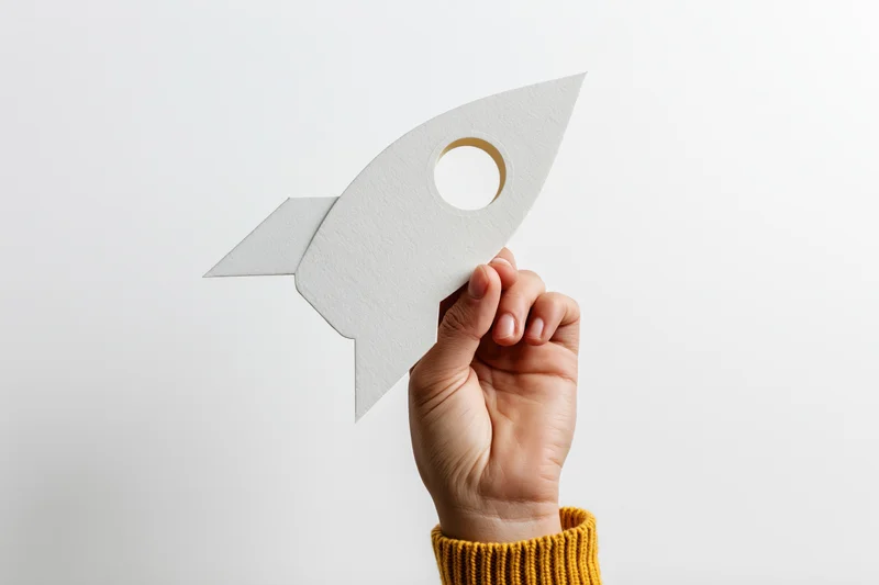 Hand holding white paper rocket cutout against plain background