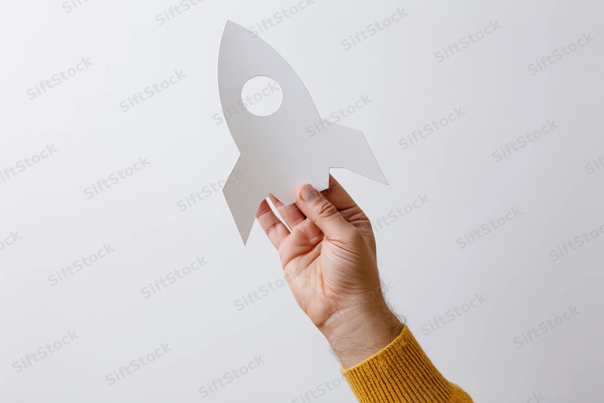 Hand holding white paper rocket cutout against plain background