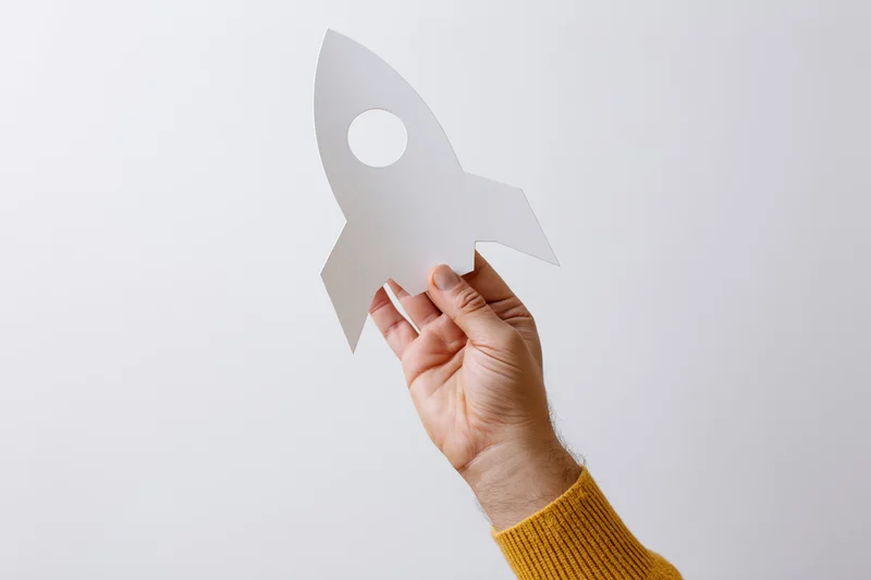 Hand holding white paper rocket cutout against plain background