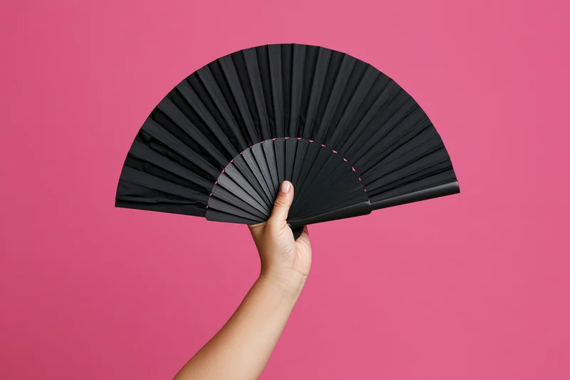 Hand holding open black folding fan against pink background