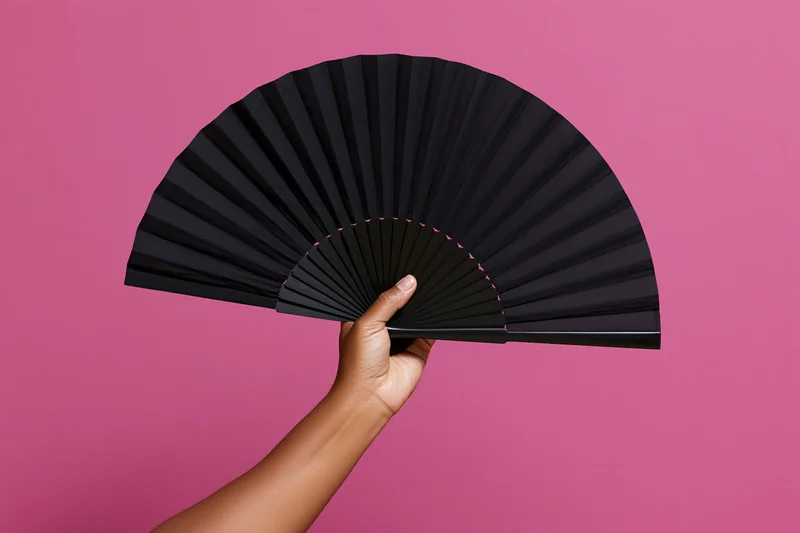 Hand holding open black folding fan against pink background