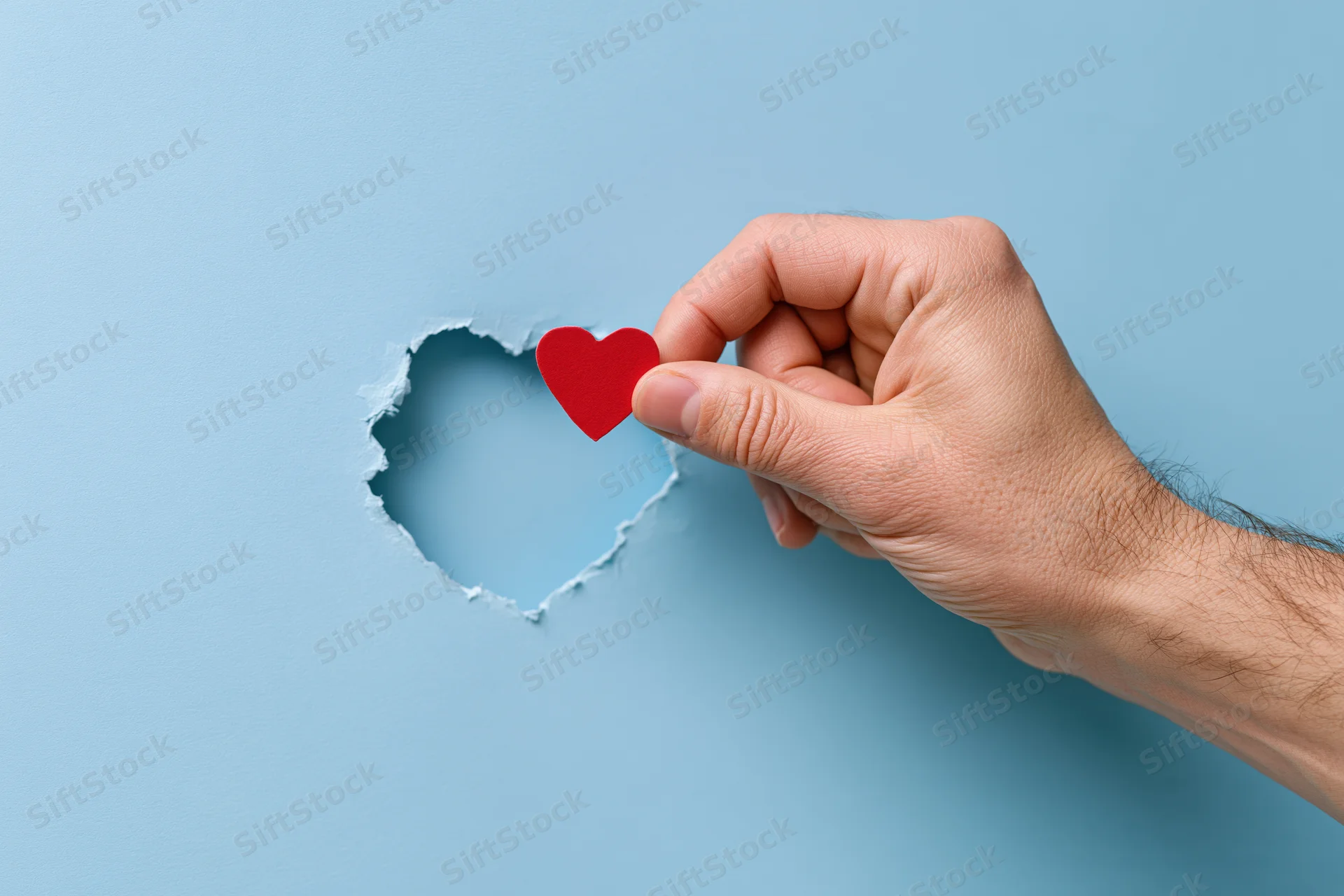 Hand holding red heart through torn hole in blue paper