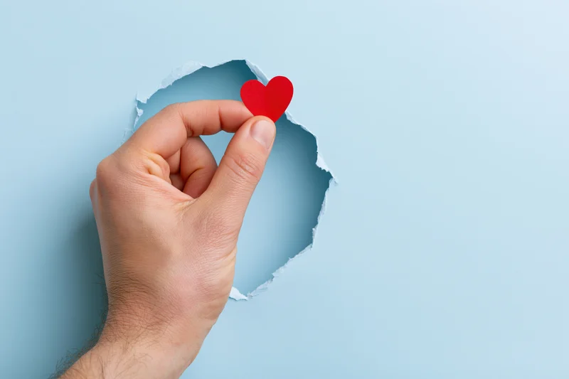Hand holding red heart through torn hole in blue paper