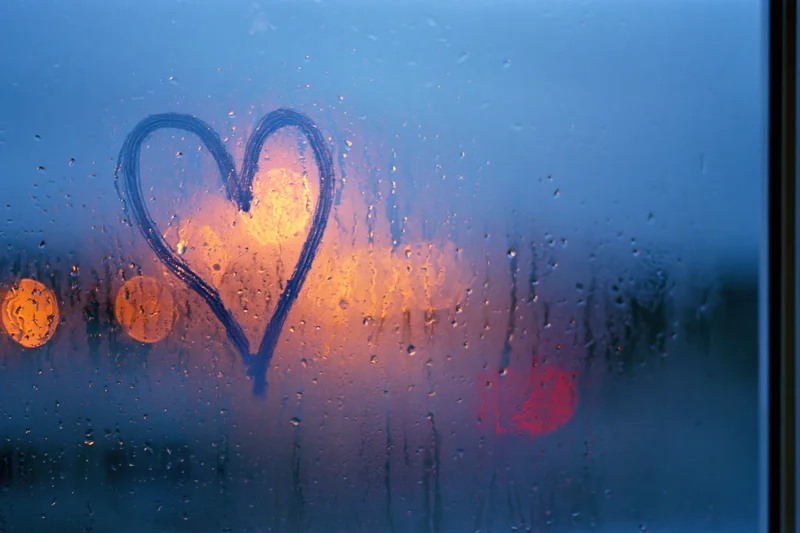 Heart drawn on fogged window with raindrops and blurred city lights at twilight