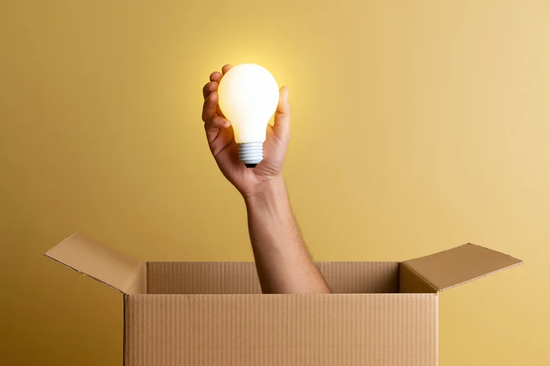 Arm reaching from cardboard box holding glowing lightbulb against yellow background