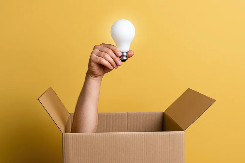 Arm reaching from cardboard box holding lightbulb against yellow background