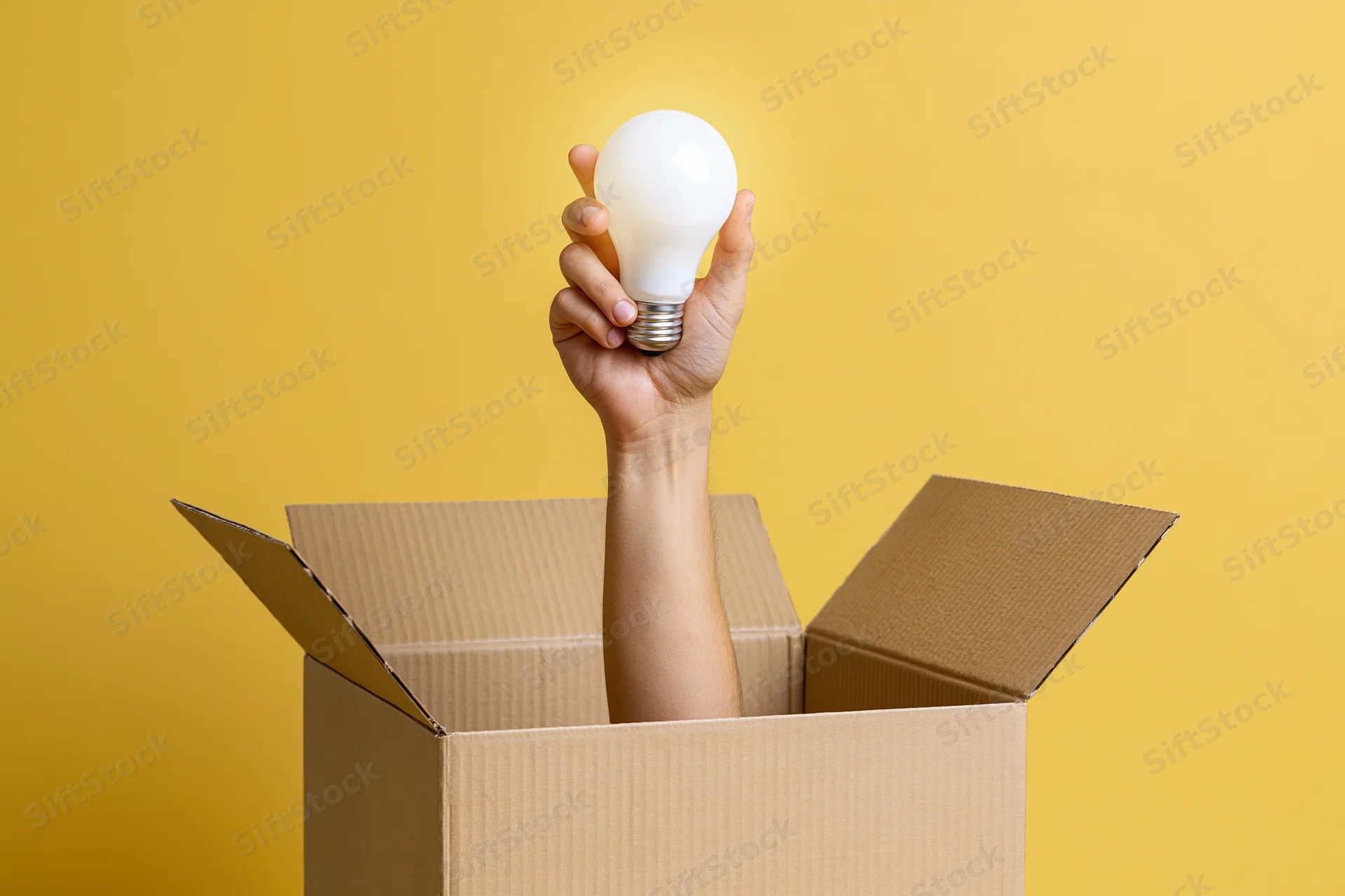 Arm reaching from cardboard box holding lightbulb against yellow background