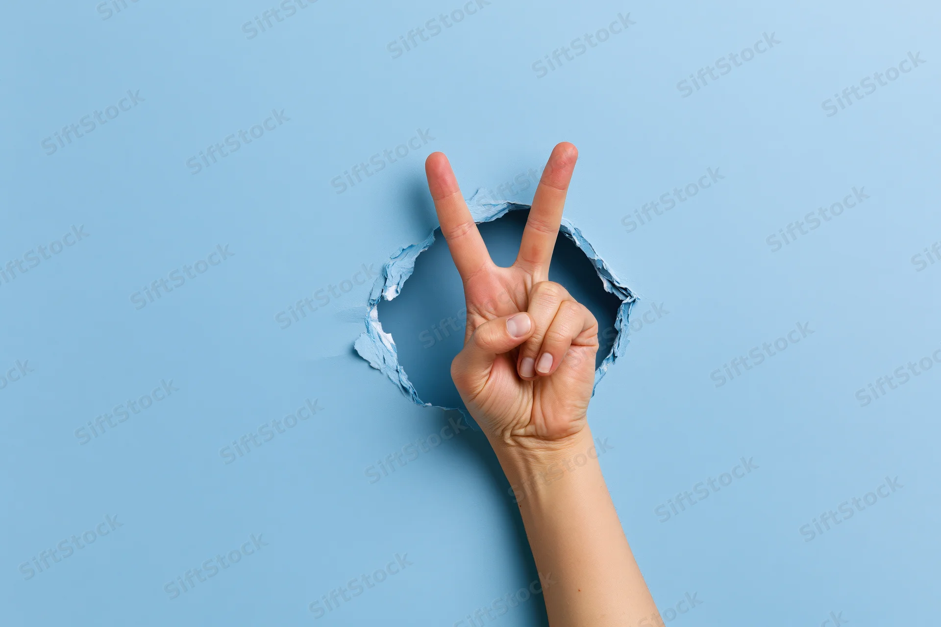 Hand making peace sign emerging through torn blue paper