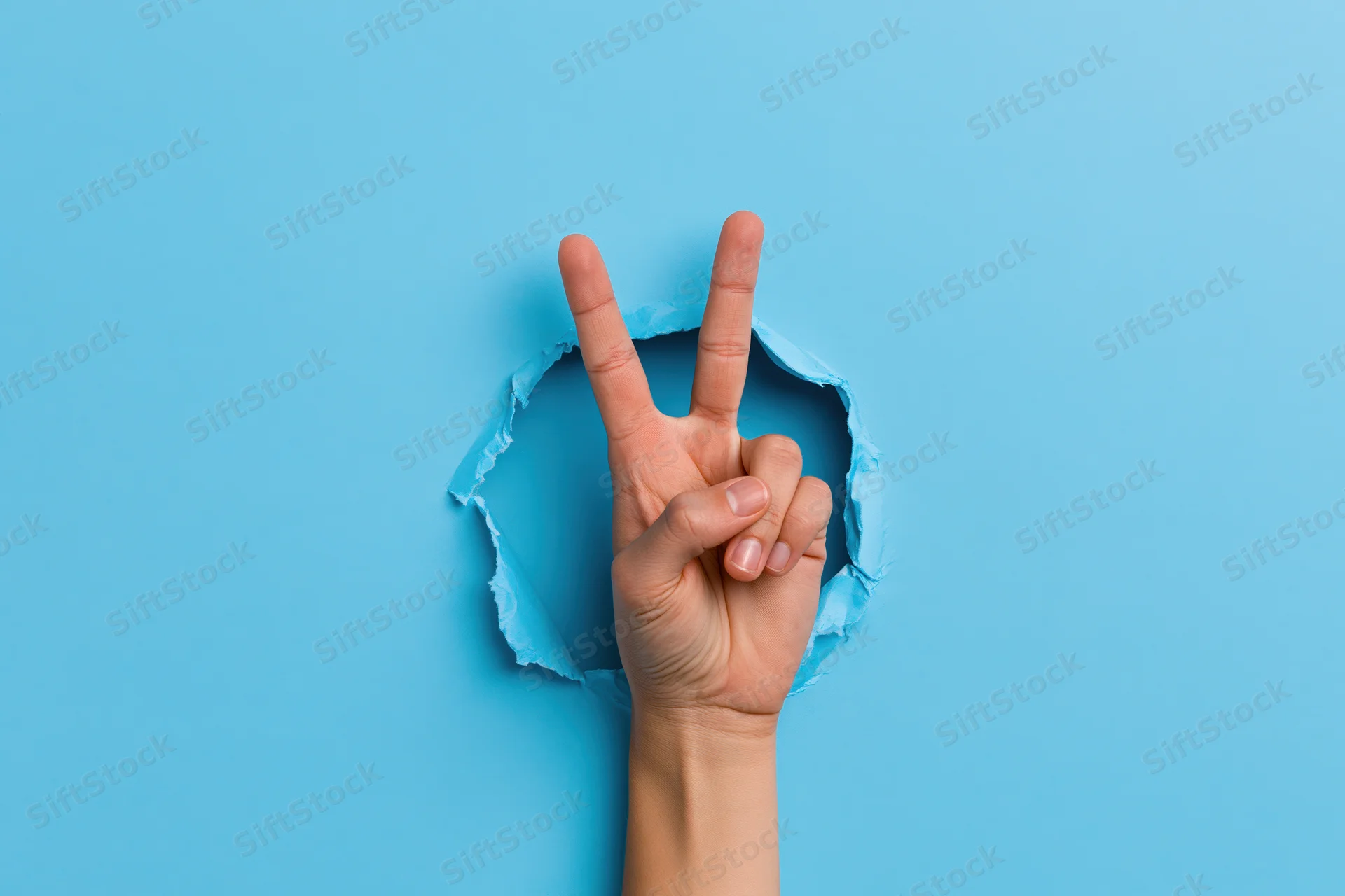 Hand making peace sign emerging through torn blue paper background