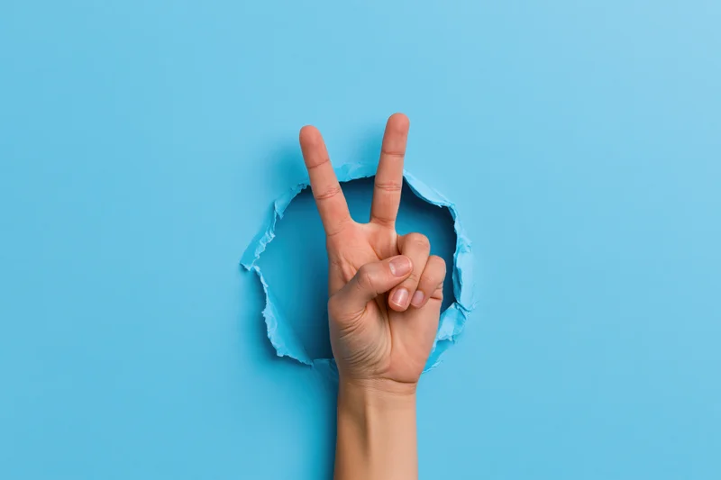 Hand making peace sign emerging through torn blue paper background