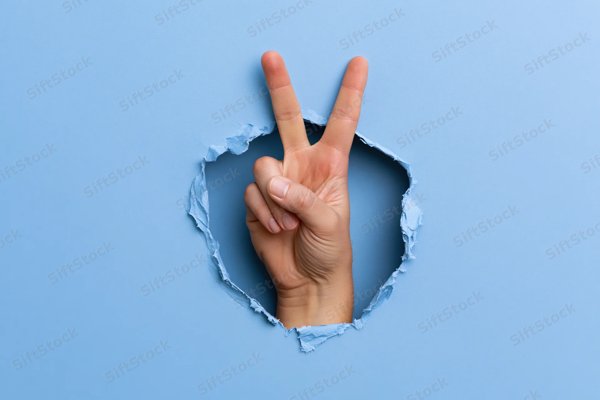 Hand making peace sign emerging through torn blue paper hole