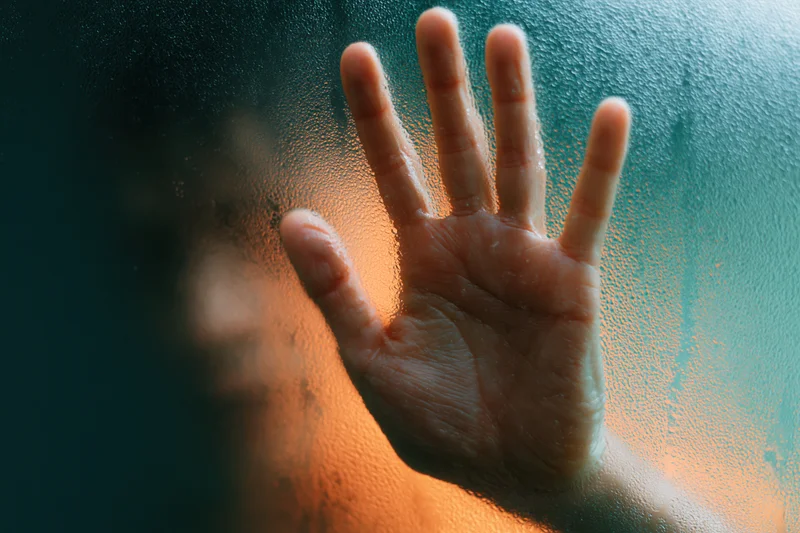 Hand pressed against wet frosted glass in a shower with warm light behind