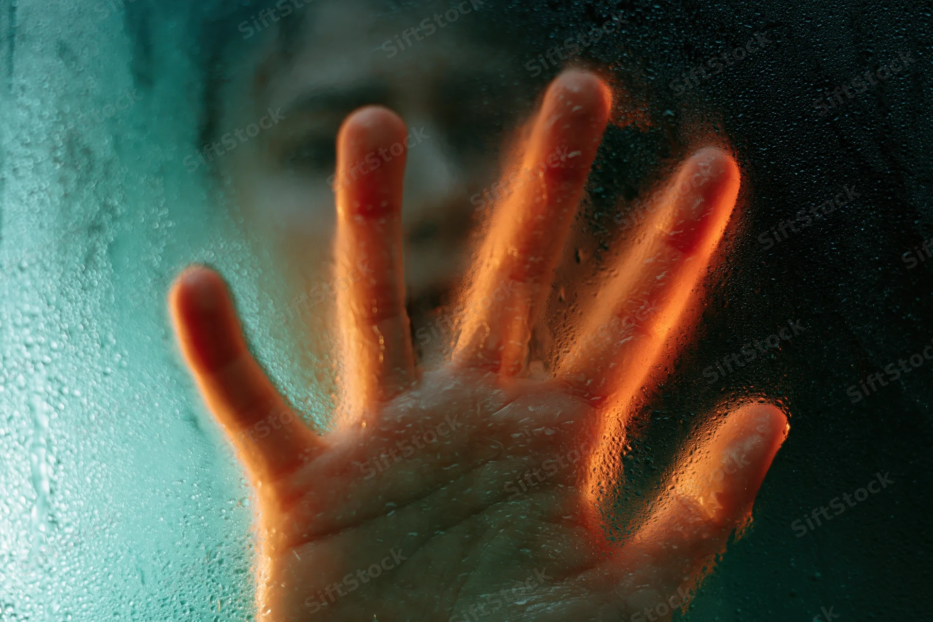 Hand pressed against wet glass with condensation and blurred face behind it