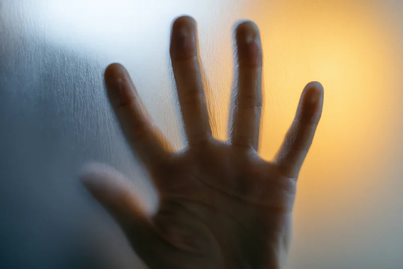 Hand pressed flat against frosted glass with fingers spread, light glowing through from behind