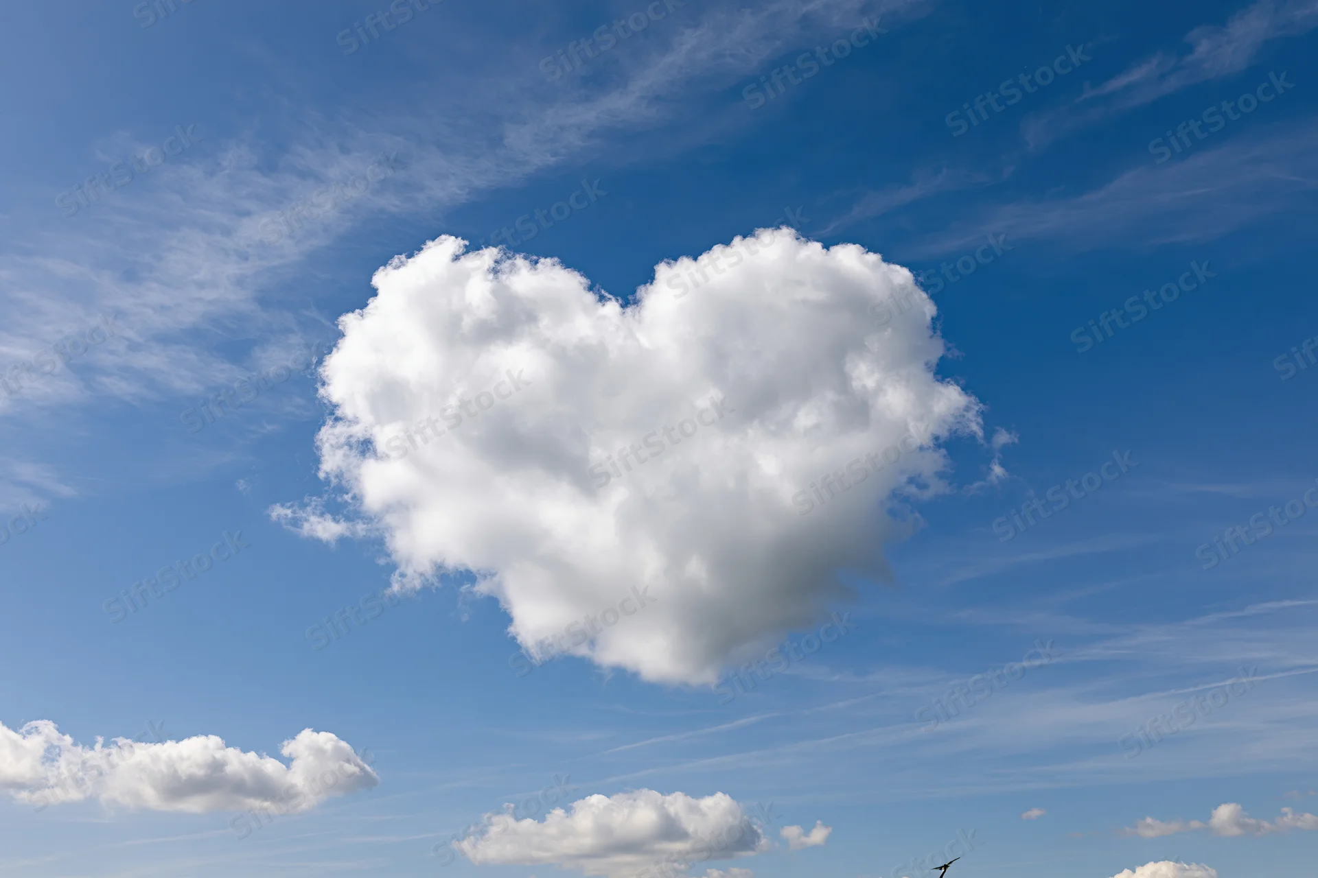 Heart-shaped cloud floating in a clear blue sky with scattered wispy clouds and a bird silhouette below