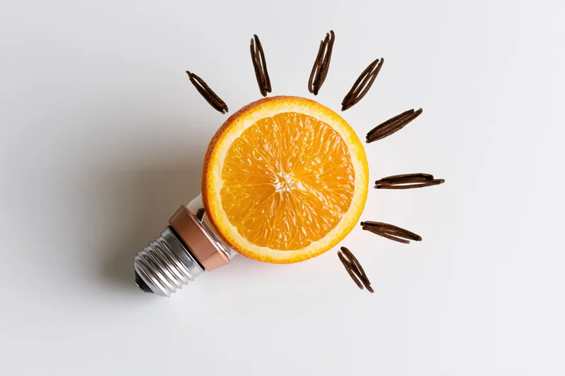 Orange slice replacing glass globe of lightbulb on white surface with cinnamon sticks as rays