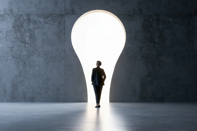 Woman in dark suit standing with back to camera facing bright lightbulb-shaped doorway in concrete room