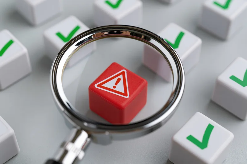 Magnifying glass focusing on a red cube with a white exclamation mark among white cubes with green checkmarks on a gray surface