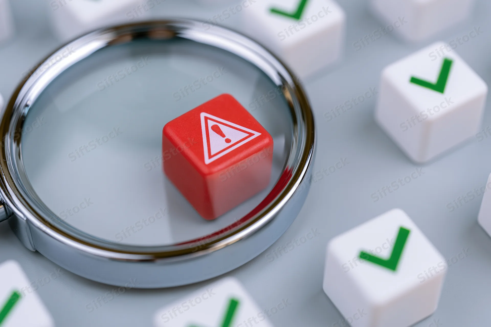 Magnifying glass focusing on a red cube with warning symbol among white cubes with green checkmarks on a gray surface