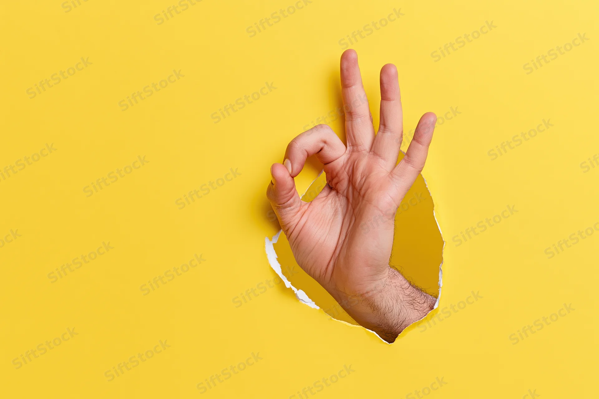 Man's hand making ok gesture emerging through torn hole in yellow paper
