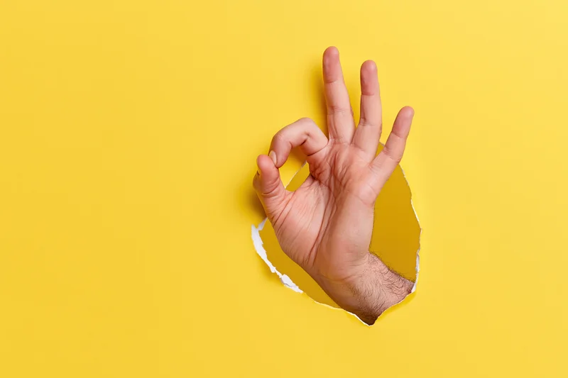 Man's hand making ok gesture emerging through torn hole in yellow paper