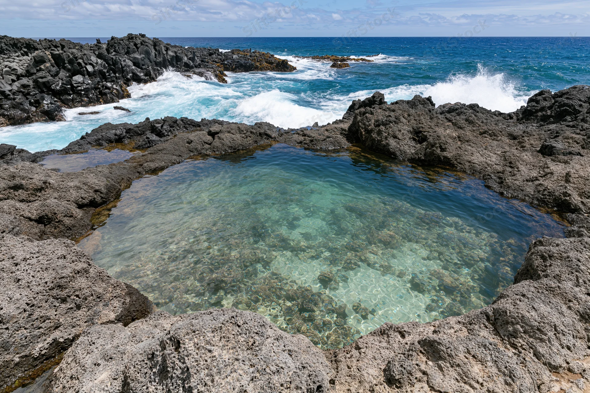 Clear turquoise water in a natural lava rock tidal pool surrounded by ocean waves and rugged coastline
