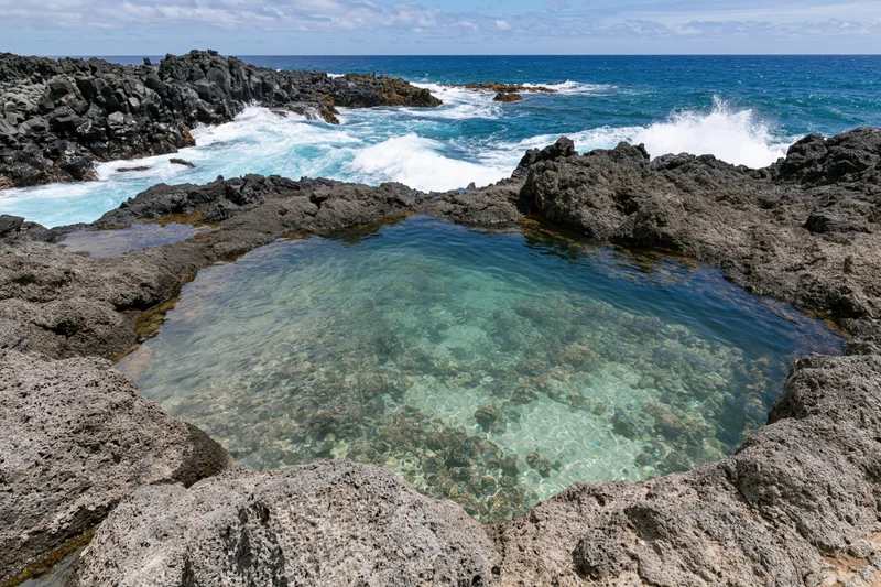 Clear turquoise water in a natural lava rock tidal pool surrounded by ocean waves and rugged coastline
