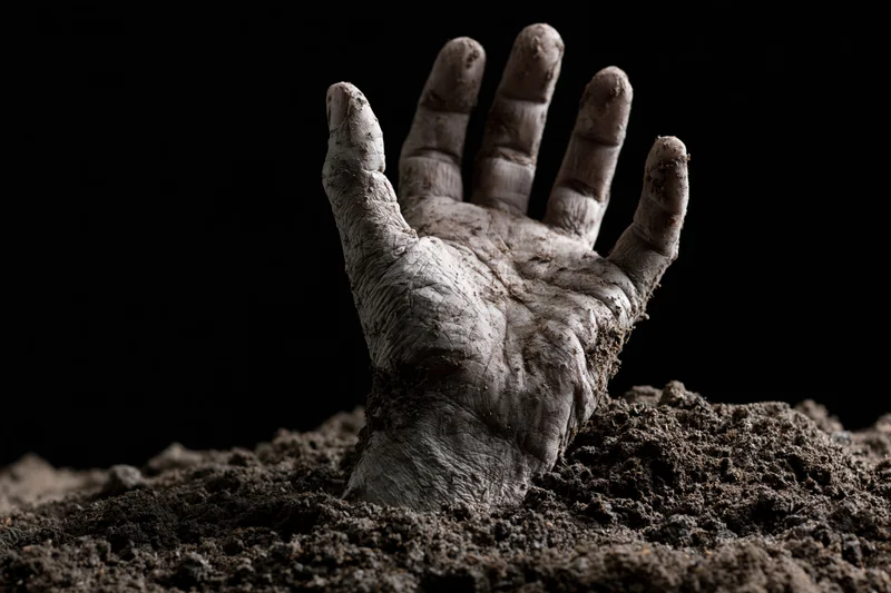Decayed hand emerging from soil with fingers splayed against dark background