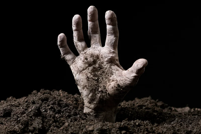 Decayed pale hand reaching up from dark soil against black background