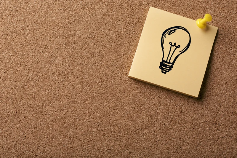 Sticky note with hand-drawn lightbulb pinned to corkboard in office workspace