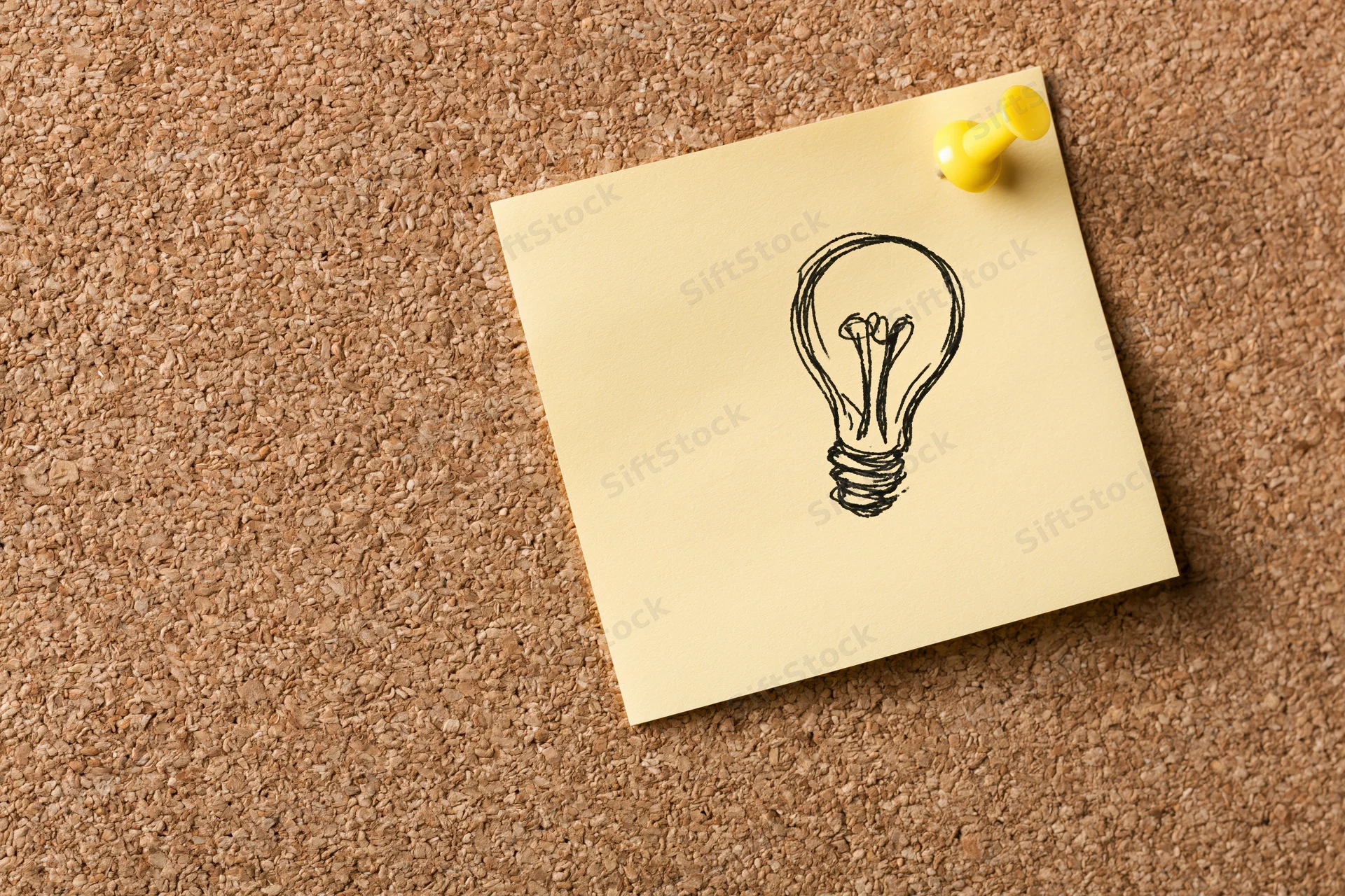Hand-drawn lightbulb sketch on a yellow sticky note pinned to a cork bulletin board
