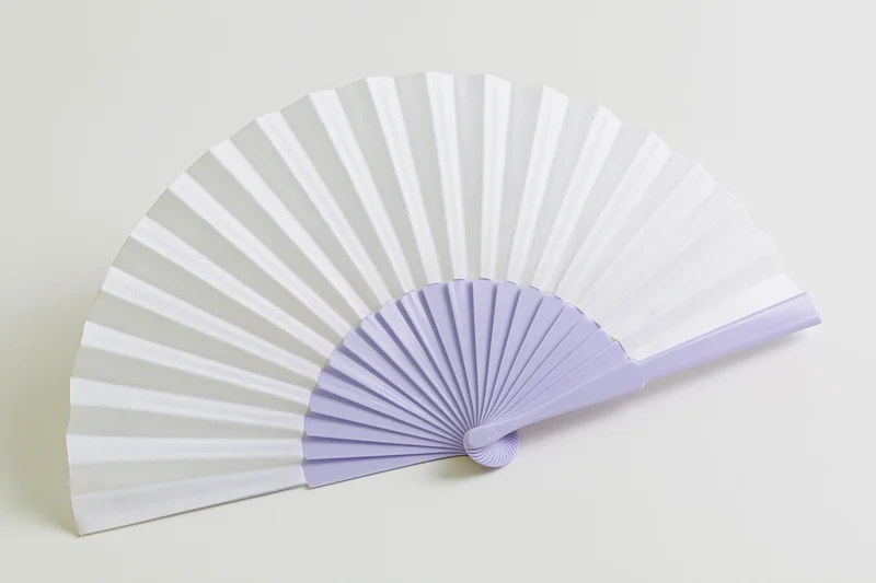 Open folding hand fan with white paper and lavender plastic frame lying flat on light beige surface