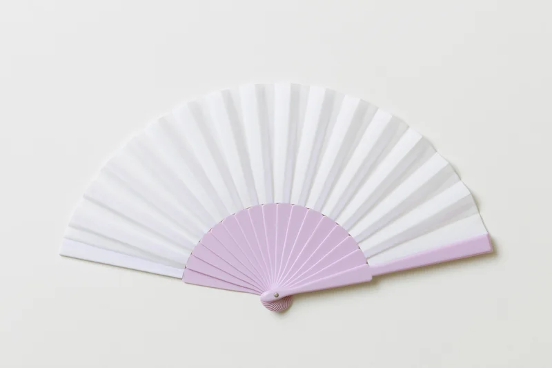 Open folding hand fan with white paper and pink plastic frame lying flat on cream background