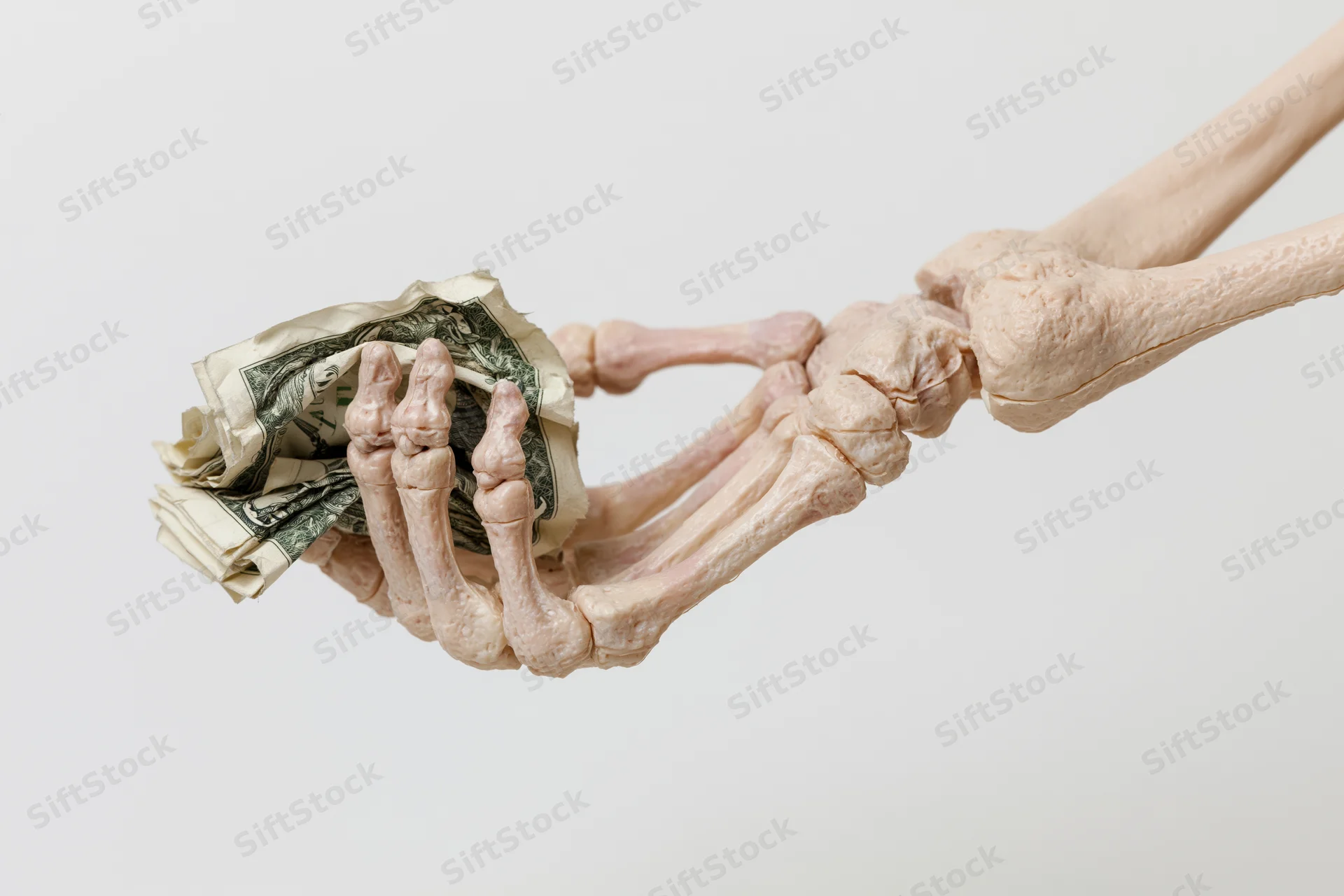 Plastic skeleton hand gripping crumpled us dollar bills against white background