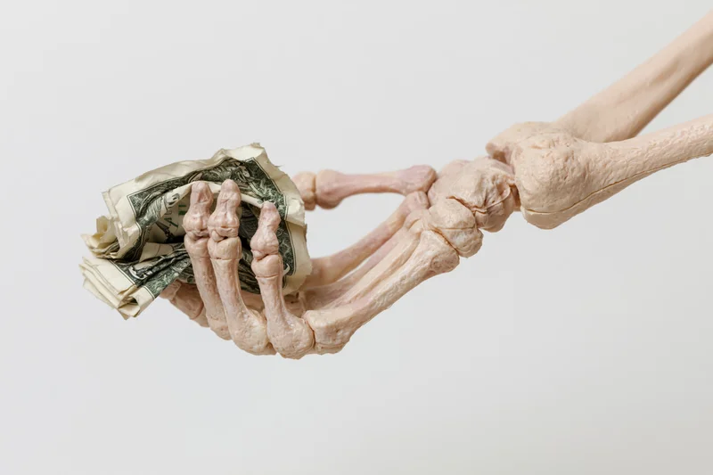 Plastic skeleton hand gripping crumpled us dollar bills against white background
