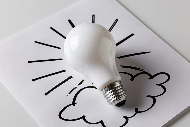 White lightbulb resting on paper with hand-drawn sun and cloud sketch