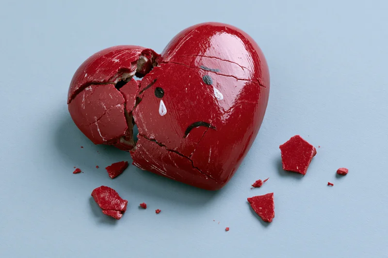 Broken red ceramic heart with painted sad crying face on blue background