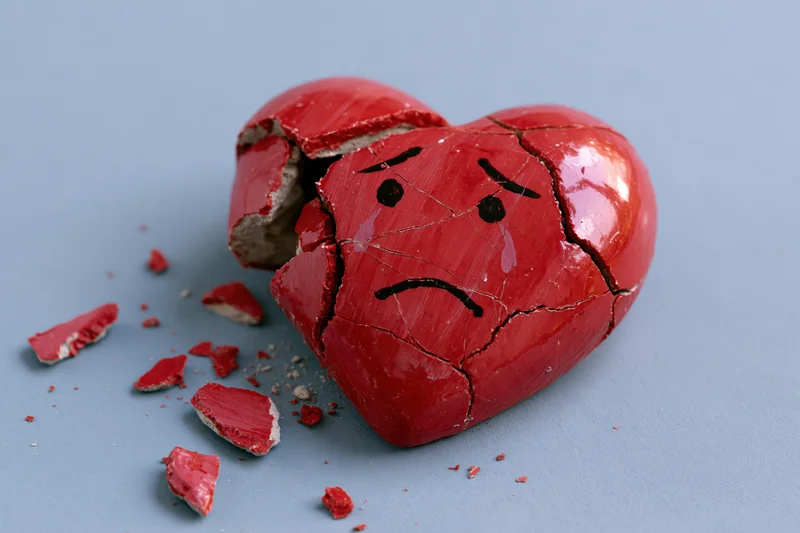 Broken red ceramic heart with sad crying face painted on it, scattered fragments on gray surface