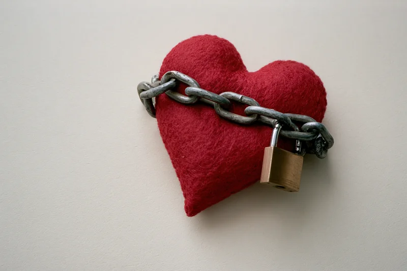 Red fabric heart wrapped with metal chain and brass padlock on white surface