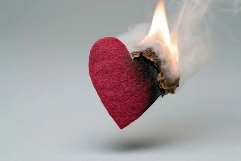 Red felt heart cutout burning with flame and smoke against light background