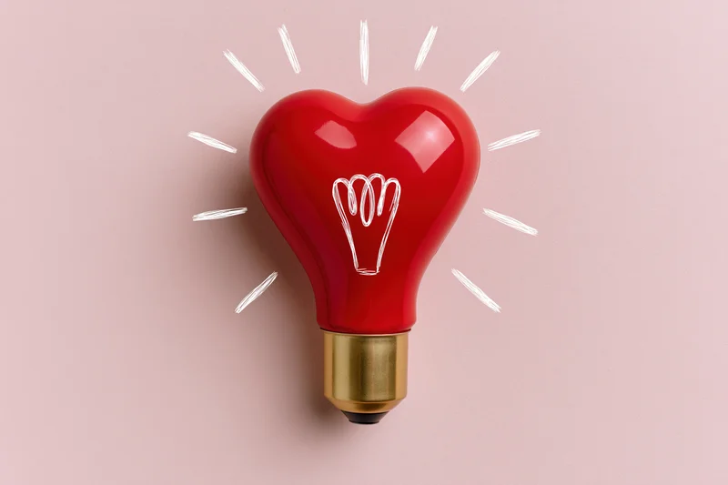 Red heart-shaped lightbulb with gold base and white drawn filament on pink background
