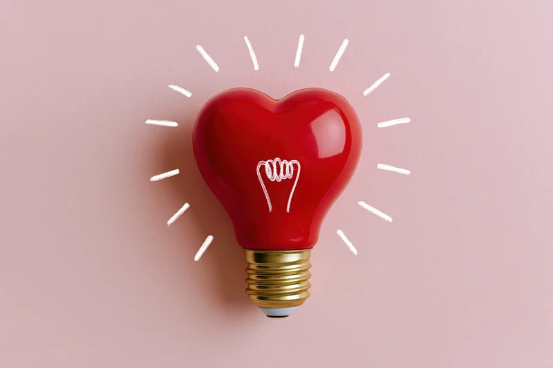 Red heart-shaped lightbulb with gold base and white glow lines on pink background