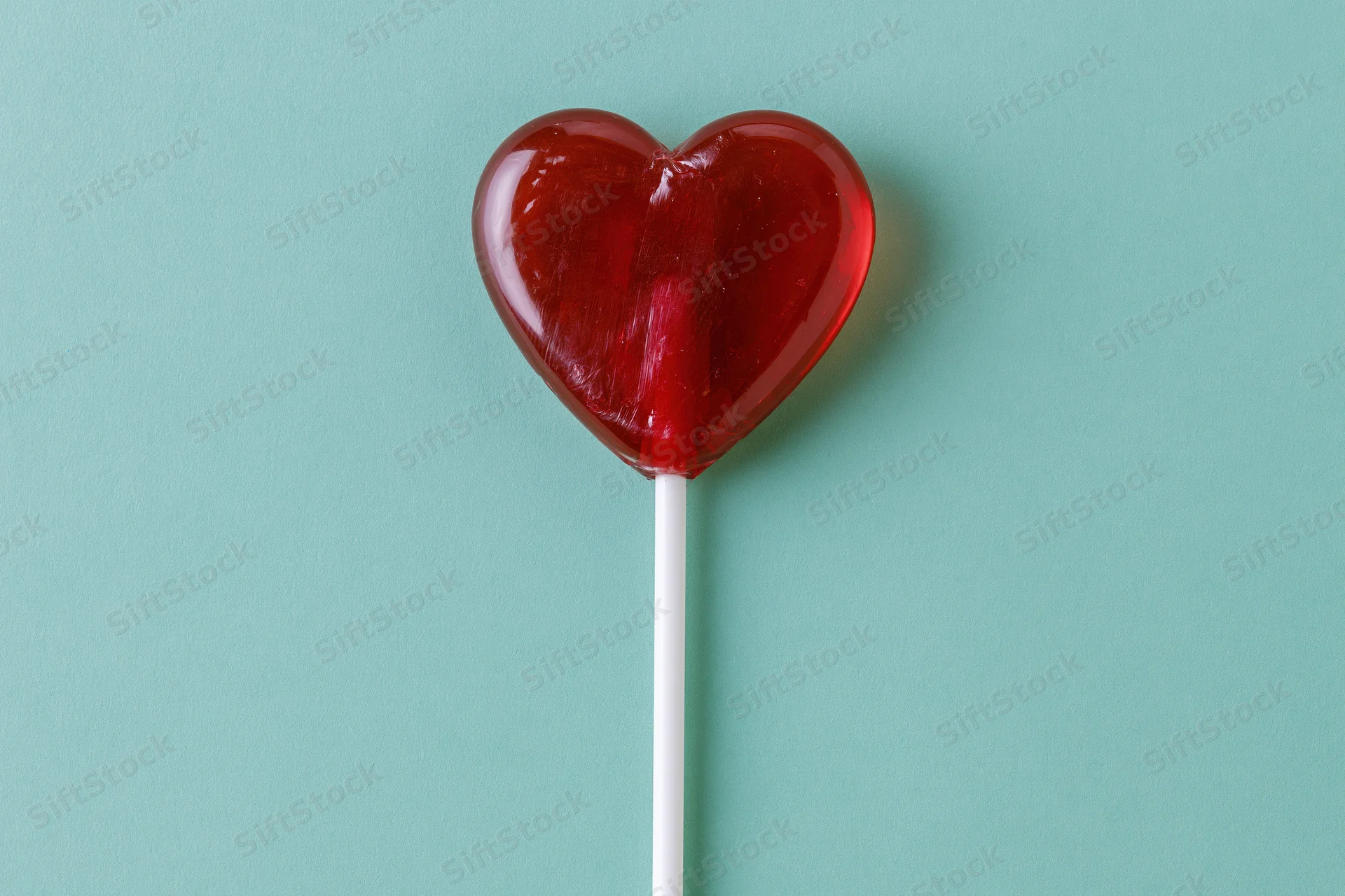 Red heart-shaped lollipop on white stick against teal background
