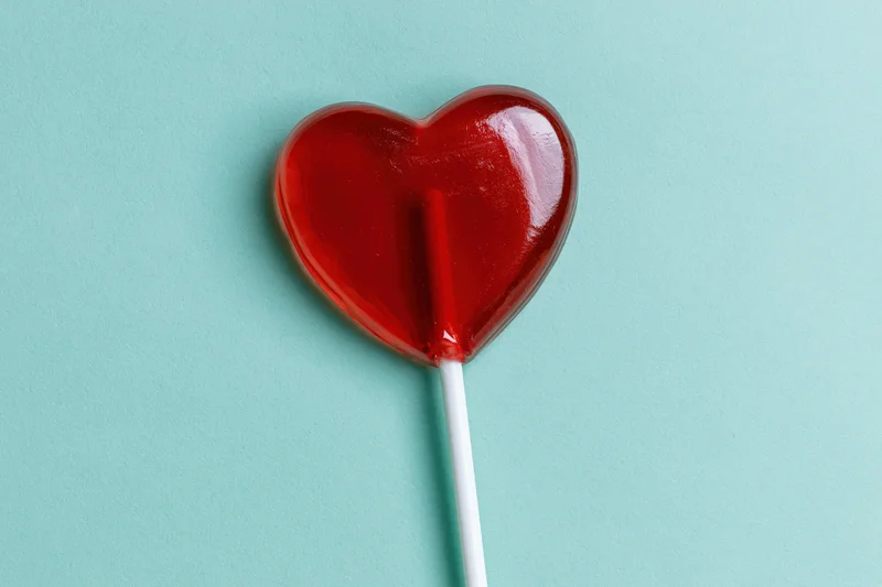 Red heart-shaped lollipop on white stick against turquoise background
