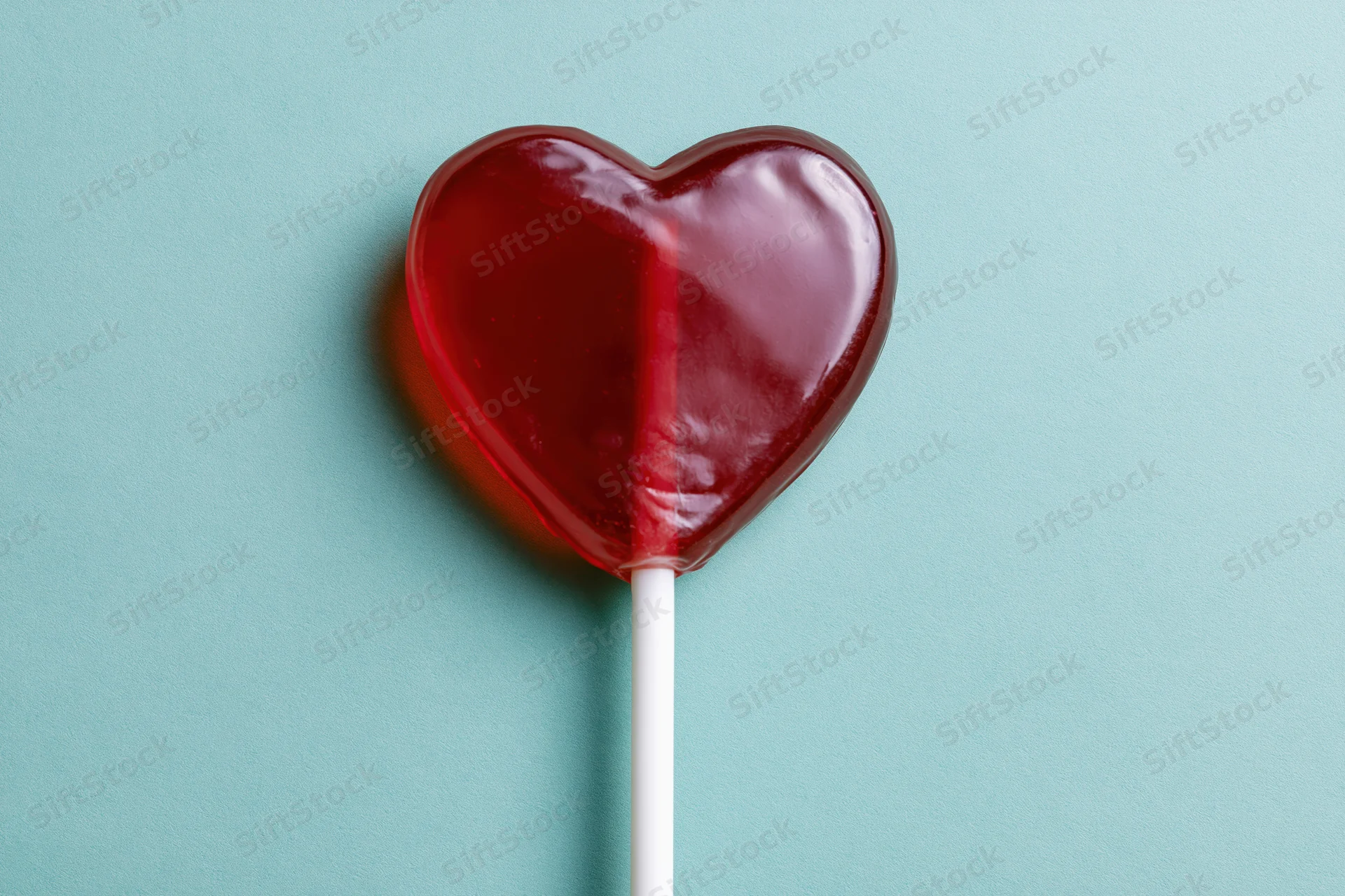 Heart-shaped red lollipop on white stick against teal background