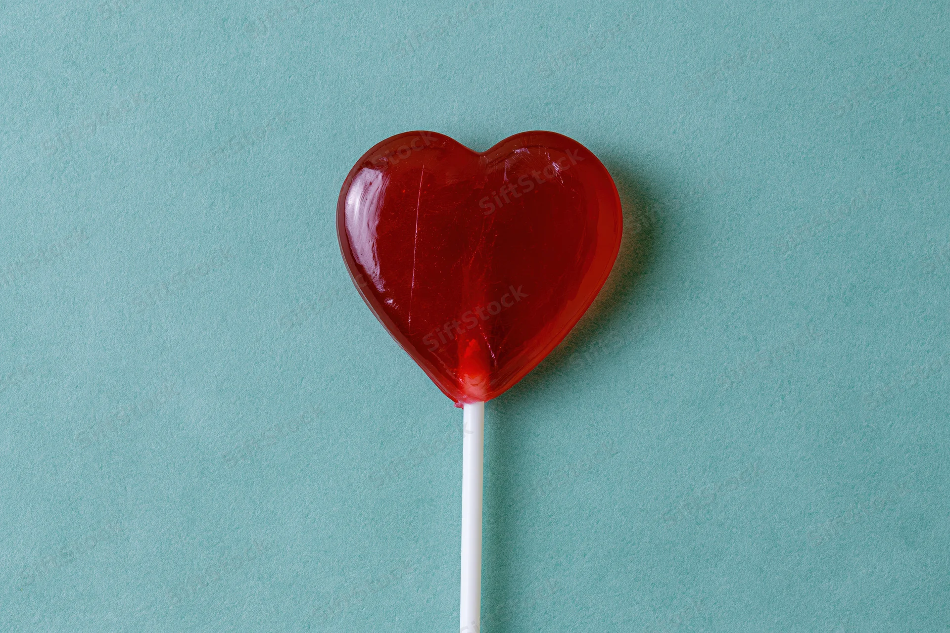 Red heart-shaped lollipop on white stick against turquoise background