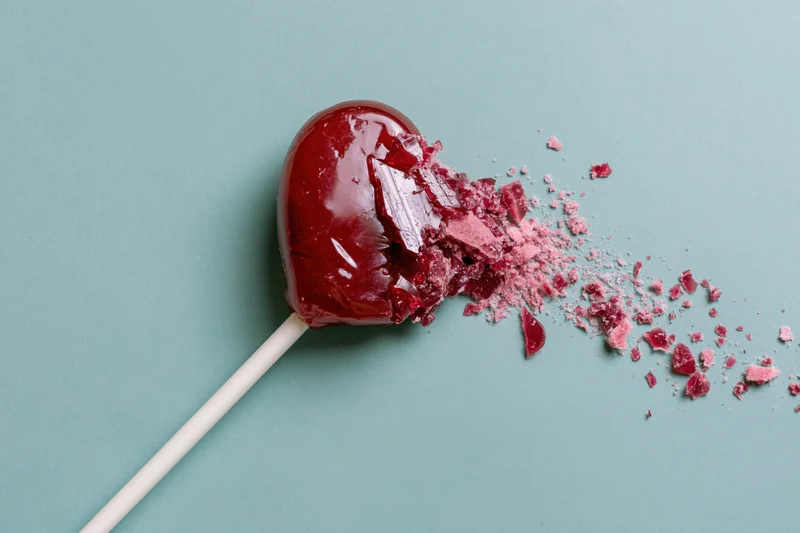 Broken red heart-shaped lollipop with shattered candy fragments on teal background