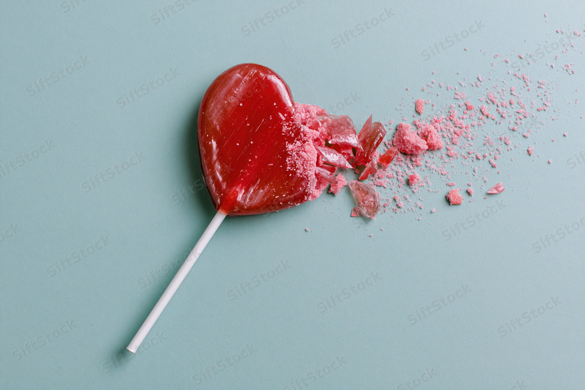 Broken red heart-shaped lollipop with shattered pieces on a pastel blue background