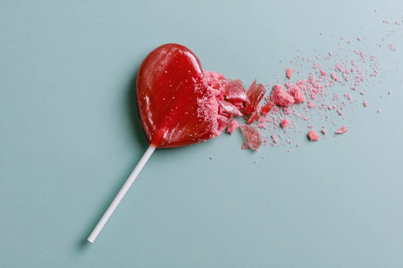 Broken red heart-shaped lollipop with shattered pieces on a pastel blue background